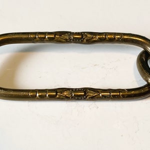 May include: Close-up of a brass chain link with a decorative pattern. The link is oval-shaped and has a smooth, polished finish.