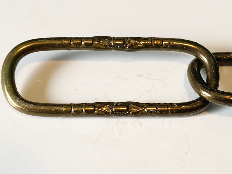 May include: Close-up of a brass chain link with a decorative pattern. The link is oval-shaped and has a smooth, polished finish.