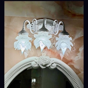 May include: A white wall-mounted light fixture featuring three downward-facing glass shades shaped like flower petals. The metal base has curved arms and decorative scrollwork, positioned above a white oval mirror frame with a carved rose detail.