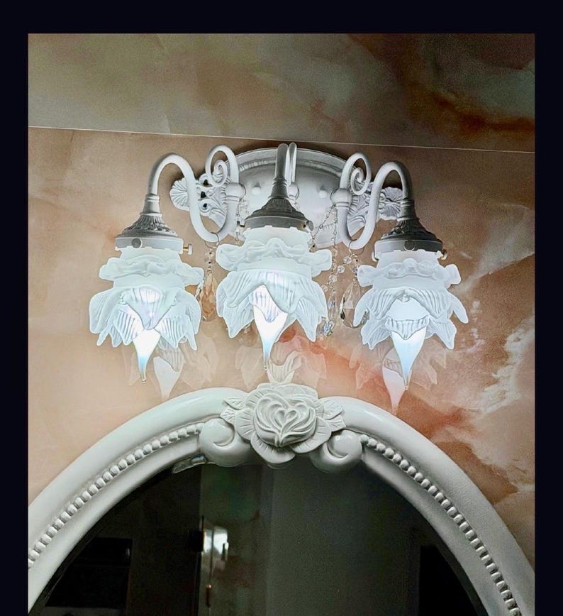 May include: A white wall-mounted light fixture featuring three downward-facing glass shades shaped like flower petals. The metal base has curved arms and decorative scrollwork, positioned above a white oval mirror frame with a carved rose detail.