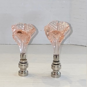 Peut inclure: Deux embouts décoratifs pour lampes. Chaque embout présente un dessus en verre rose en forme de fleur et une base en métal argenté. Les dessus de fleurs ont un design à facettes et les bases ont un aspect superposé. Les embouts sont posés sur un fond blanc.