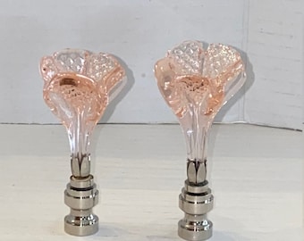 May include: Two decorative lamp finials. Each finial features a pink, flower-shaped glass top and a silver-colored metal base. The flower tops have a faceted design, and the bases have a layered appearance. The finials are set against a white background.