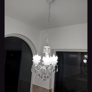 May include: A white metal chandelier featuring three light fixtures shaped like open flower petals. The fixture is suspended by a matching white chain and is adorned with numerous clear, faceted crystal teardrop pendants hanging from the arms.