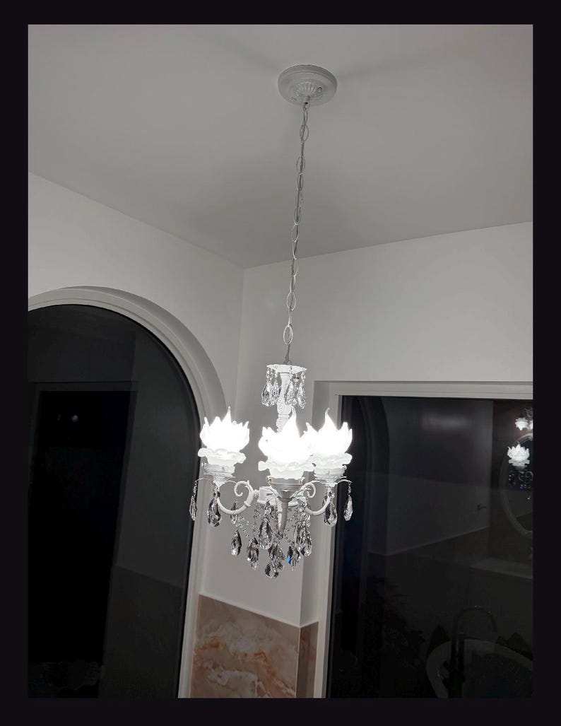 May include: A white metal chandelier featuring three light fixtures shaped like open flower petals. The fixture is suspended by a matching white chain and is adorned with numerous clear, faceted crystal teardrop pendants hanging from the arms.