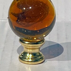 May include: A gold lamp finial with a clear amber glass sphere. The glass sphere has a swirling pattern inside.