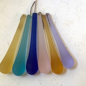 Op de afbeelding: Zes glazen ornamenten in de vorm van een traan in verschillende pastelkleuren, waaronder geel, blauw, turquoise, paars en roze, hangend aan een draad.