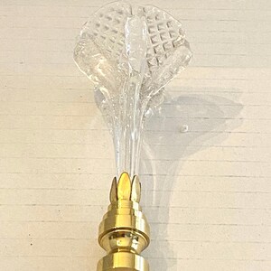 May include: A clear glass lamp finial with a textured, flower-like top and a gold-colored metal base. The finial is designed to be screwed onto a lamp. The glass top has a diamond pattern.