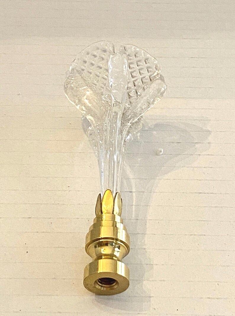 May include: A clear glass lamp finial with a textured, flower-like top and a gold-colored metal base. The finial is designed to be screwed onto a lamp. The glass top has a diamond pattern.