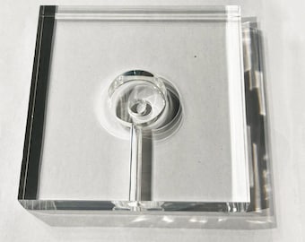 May include: A clear, square acrylic display block featuring a centered circular indentation and a vertical channel extending to the bottom edge. The transparent material has polished edges and creates distinct shadows on the surface below.