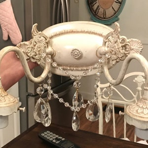 May include: A two-light wall sconce featuring an off-white, distressed finish with ornate scrollwork and decorative molding. The fixture includes two curved arms and is adorned with clear, faceted crystal teardrop pendants and beaded chains.