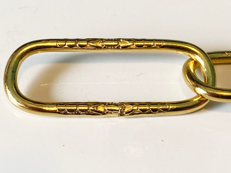 May include: Close-up of a gold chain link with a decorative pattern. The link is oval-shaped and has a smooth, polished finish.