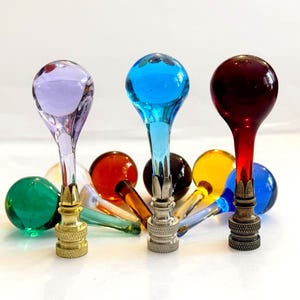 May include: A collection of lamp finials featuring spherical glass tops in various colors including lavender, blue, red, green, yellow, and amber. Each finial has a metal base with a decorative collar, available in gold, silver, and bronze finishes.