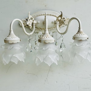 May include: A three-light wall sconce featuring an off-white metal frame with ornate scrollwork and an oval backplate. Three frosted glass shades shaped like flower petals are suspended from curved arms, accented by clear hanging crystal drops.