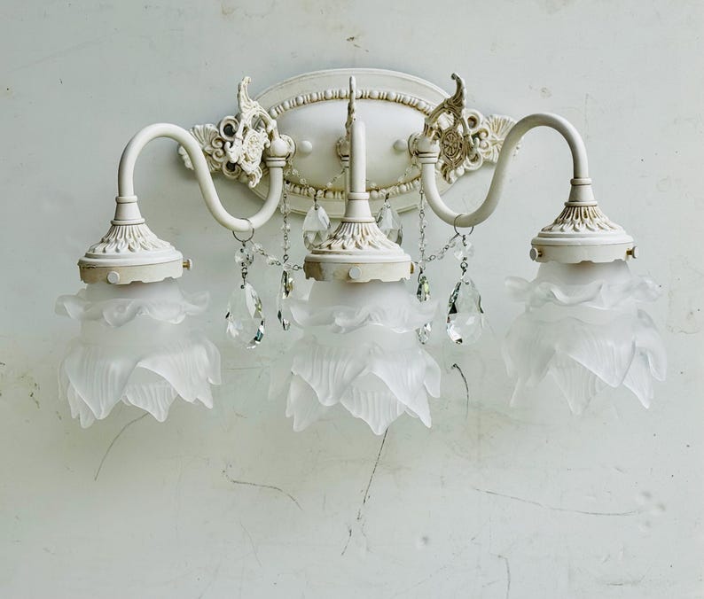 May include: A three-light wall sconce featuring an off-white metal frame with ornate scrollwork and an oval backplate. Three frosted glass shades shaped like flower petals are suspended from curved arms, accented by clear hanging crystal drops.