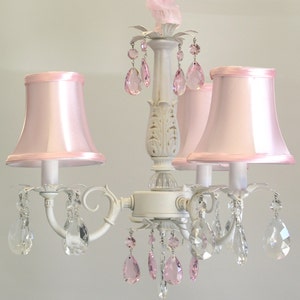 May include: A white chandelier with pink fabric lampshades and crystal teardrop accents. The chandelier has a traditional design with a central column and three arms.