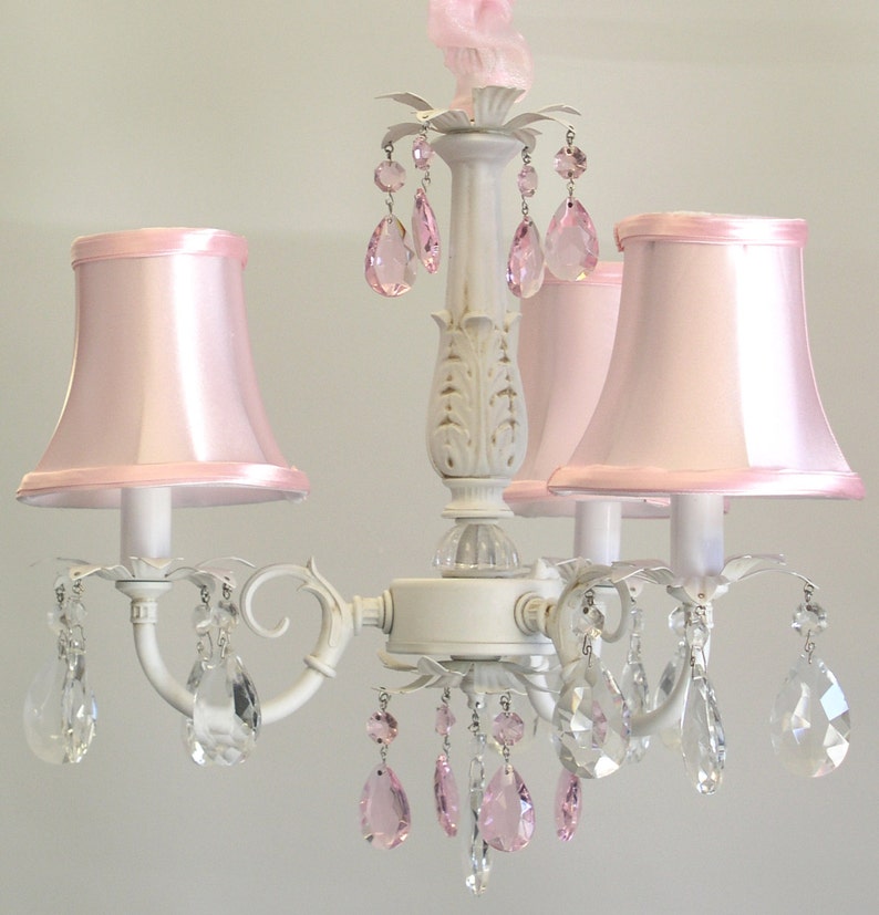 May include: A white chandelier with pink fabric lampshades and crystal teardrop accents. The chandelier has a traditional design with a central column and three arms.