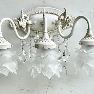 May include: A three-light wall sconce featuring a white metal base with ornate detailing. Three frosted glass shades shaped like flower petals are suspended from curved arms. Clear crystal teardrop accents hang between the light fixtures.