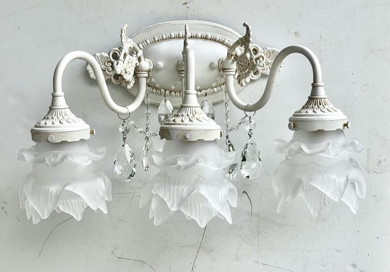 May include: A three-light wall sconce featuring a white metal base with ornate detailing. Three frosted glass shades shaped like flower petals are suspended from curved arms. Clear crystal teardrop accents hang between the light fixtures.