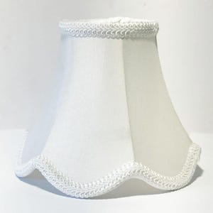 Könnte beinhalten: Weißer Lampenschirm mit gewelltem Rand und dekorativer Verzierung. Der Lampenschirm hat eine klassische Glockenform und besteht aus einem glatten, undurchsichtigen Stoff. Die Verzierung ist ein zartes, weißes, gewebtes Design.