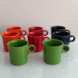 Puede incluir: Un conjunto de tazas de cerámica en varios colores, incluyendo naranja, rojo, verde y azul marino. Cada taza tiene un asa redondeada y un diseño sencillo y clásico. Las tazas están dispuestas sobre una superficie blanca.