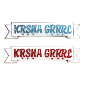 May include: Two white enamel pins with a banner design. The top pin has the text "KRISNA GRRRR" in blue glitter. The bottom pin has the text "KRISNA GRRRR" in red glitter.