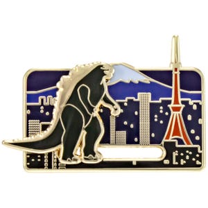 Könnte beinhalten: Goldfarbene Anstecknadel mit einer schwarzen Godzilla-Figur vor einer nächtlichen Stadtlandschaft mit einem roten Turm und einem Berg. Die Anstecknadel hat eine rechteckige Form mit einem goldenen Rand und einem Nadelverschluss.