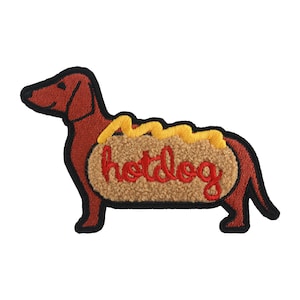 Może przedstawiać: Haftowana naszywka przedstawiająca jamnika w kształcie hot doga. Pies jest brązowy z czarnym obrysem, a bułka do hot doga jest beżowa z żółtą musztardą. Słowo "hotdog" jest w czerwonym skrypcie.
