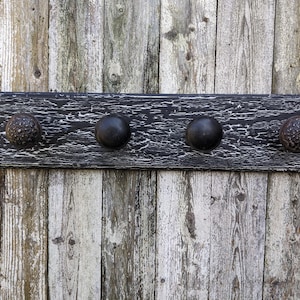 Op de afbeelding: Een zwarte houten kapstok met vier zwarte metalen haken. De kapstok heeft een verweerde afwerking en is gemonteerd op een verweerde houten muur.