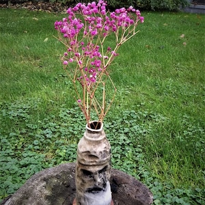 Puede incluir: Un jarrón de cerámica con un diseño texturizado con forma de cara, que contiene un ramo de flores rosas. El jarrón está sentado sobre un tocón de árbol.