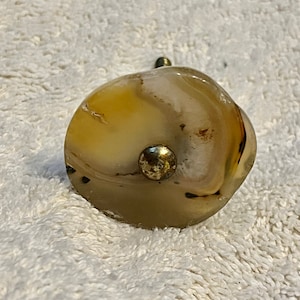 May include: A decorative cabinet knob made of polished agate stone. The knob features a mix of beige, brown, and white colors with a small, round, gold-colored metal accent. The knob has a threaded metal post for attachment.