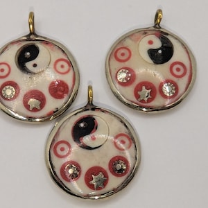 May include: Three circular pendants featuring a central black and white yin-yang symbol on a cream-colored background. The pendants are accented with red circular patterns and small metallic star and flower-shaped embellishments, all encased in a gold-toned metal frame with a top loop for hanging.