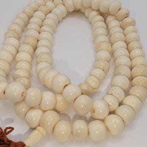 May include: A strand of off-white, disc-shaped beads arranged in a circular loop. The beads have a smooth, matte texture and are strung together, terminating at one end with a small, carved bead and a brown, multi-stranded fabric tassel.
