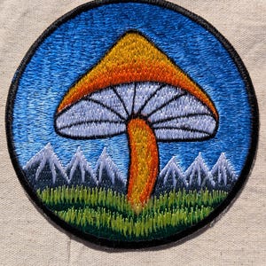 May include: A circular embroidered patch featuring a central mushroom with an orange cap and yellow stem. The mushroom is set against a blue sky background with white-capped grey mountains and a green grassy field at the base, all bordered by black.
