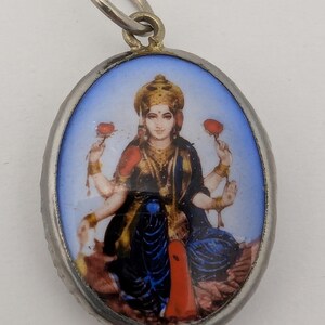 May include: An oval-shaped pendant featuring a miniature painting of a four-armed deity with a golden crown, wearing dark blue and red garments, holding red lotus flowers in the upper hands, set within a silver-toned metal frame with a jump ring.