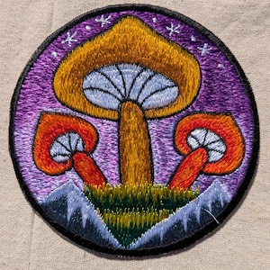 May include: A circular embroidered patch featuring three mushrooms with gold and orange caps and light blue gills. The mushrooms are set against a purple background with white star-like symbols, rising from a patch of green grass and dark blue mountains.