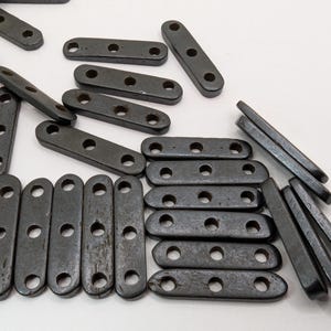 May include: A collection of dark gray, rectangular construction pieces, each featuring three circular holes arranged in a line. These flat, elongated components are scattered and organized in various orientations, showcasing a smooth, uniform finish.