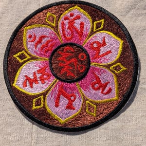 May include: A circular embroidered patch featuring a brown background with a central pink lotus flower design. Each of the six petals contains a red symbol, and the center circle displays additional red script. The design is outlined in yellow.