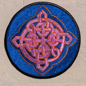 May include: A circular embroidered patch featuring a vibrant blue background with a central intricate knotwork design in shades of light purple and pink, outlined in orange thread, and finished with a solid black stitched border edge.