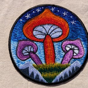 May include: A circular embroidered patch featuring a large orange mushroom in the center flanked by two smaller purple mushrooms. The background depicts a blue sky with white stars above green grass and jagged gray mountain peaks at the base.