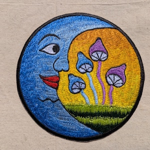 May include: A circular embroidered patch featuring a blue crescent moon with a face, red lips, and a white eye. The moon overlaps a yellow sun background containing four small mushrooms with purple and blue caps on green grass.