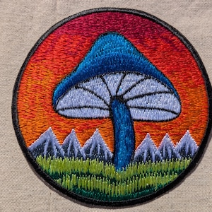 May include: A circular embroidered patch featuring a blue mushroom with a white underside, set against a vibrant orange and red sunset sky. Below the mushroom, a row of light purple mountain peaks rises from a field of green grass.