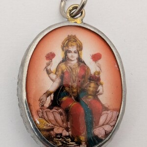 May include: An oval-shaped pendant featuring a miniature painting of a four-armed deity seated on a lotus flower. The figure wears red and gold clothing, holds red flowers in two hands, and is set within a simple silver-toned metal frame with a bail.