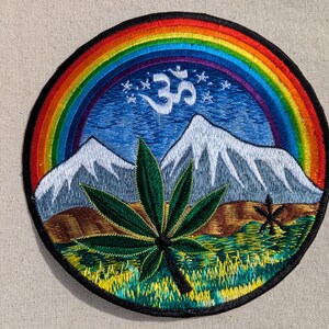 May include: A circular embroidered patch featuring a vibrant rainbow arching over snow-capped mountains and a blue sky with a white Om symbol and small stars. In the foreground, a green cannabis leaf sits above a textured yellow and green field.