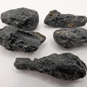 May include: Five irregular, dark black tektite stones with pitted, textured surfaces. Each piece displays a unique, rugged shape with some areas showing a smooth, glassy luster. These natural mineral specimens are arranged in a loose cluster.