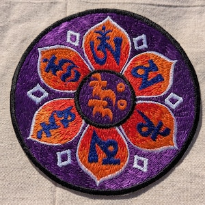 May include: A circular embroidered patch featuring a purple background with a central orange lotus flower design. Each of the six orange petals contains a blue embroidered symbol, and the center circle features orange symbols on a purple field.
