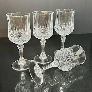 May include: Four clear crystal wine glasses with diamond-cut patterns. Three glasses stand upright, while one lies on its side. The glasses have a classic design with a round base and a detailed stem.