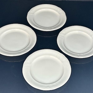 May include: Four white ceramic plates with a subtle, embossed swirl design around the rim. The plates are arranged on a dark blue surface, showcasing their clean, minimalist aesthetic. Ideal for serving meals.