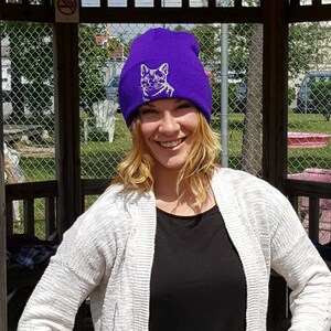 May include: A purple beanie with a white embroidered cat design. The beanie is being worn by a person with long blonde hair.