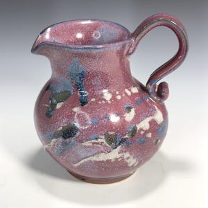 Op de afbeelding: Handgemaakte keramische kan met een afgeronde vorm en een gebogen handvat. De kan is voornamelijk dieproze van kleur, bespikkeld met witte, blauwe en zwarte accenten. Hij heeft een schenktuit en is geschikt voor het serveren van dranken.