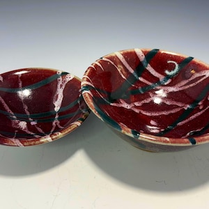 May include: Two ceramic bowls with a deep red glaze. The bowls have a glossy finish and are decorated with a pattern of white and dark green lines. The bowls are oval-shaped and appear to be handmade.